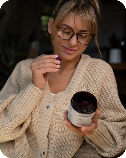 Woman in cream knit cardigan and glasses holding an open jar of purple gummy supplements and lifting one toward her mouth.