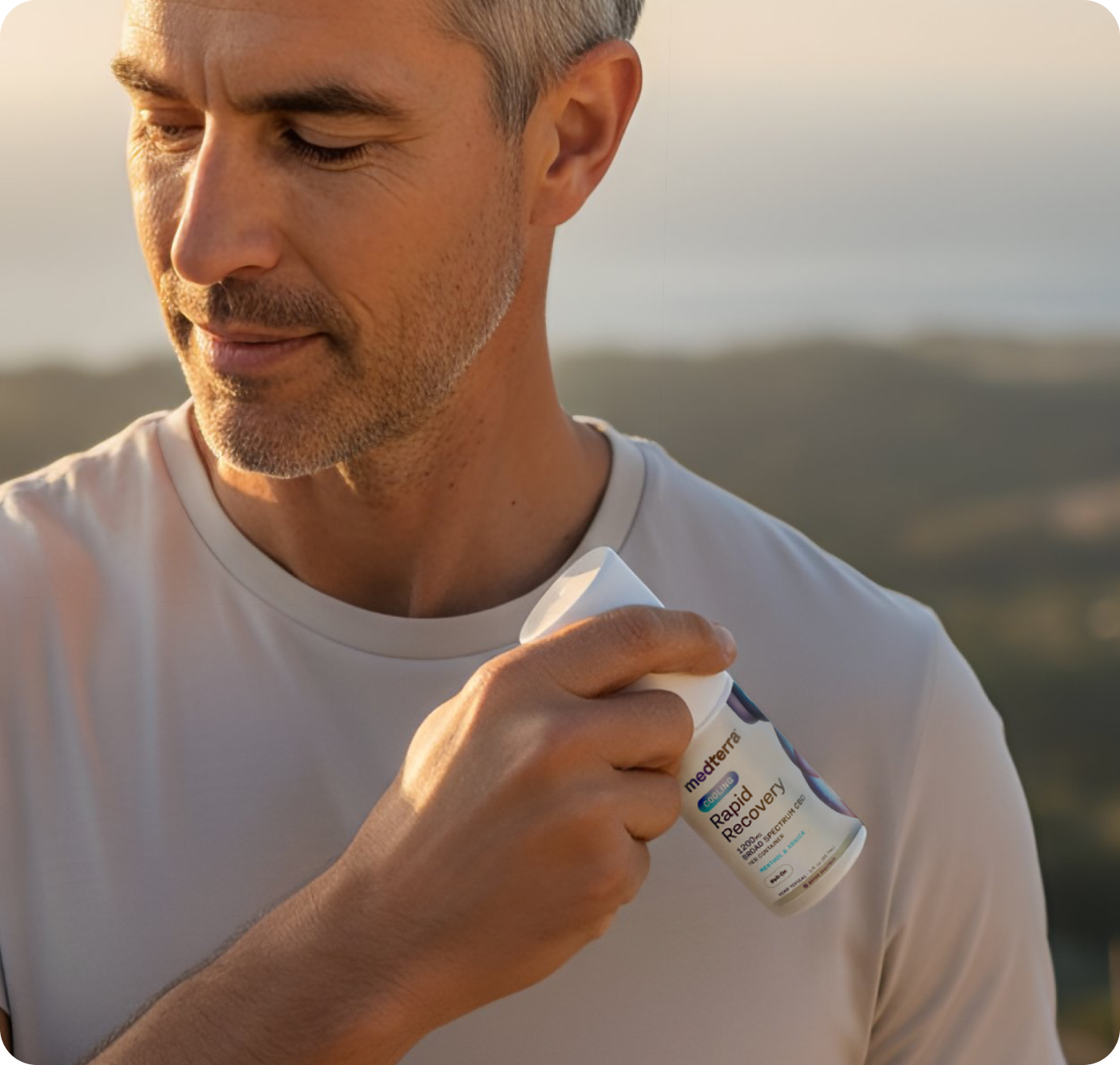 A man in a light beige shirt holds a bottle of Medterra Rapid Recovery Cooling 1200mg CBD Roll On near his shoulder, suggesting fast muscle relief, while standing outdoors with blurred natural scenery in the background.