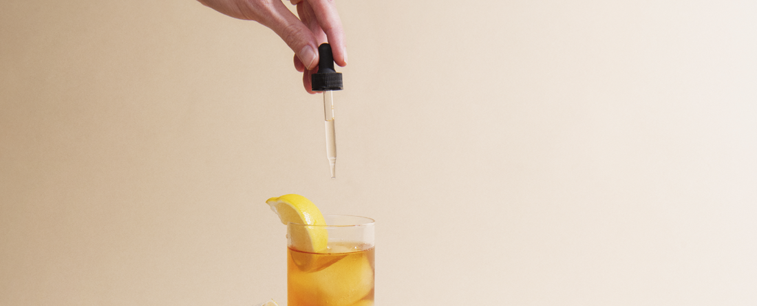 Hand holding a dropper above a glass of iced tea with a lemon slice on the rim.