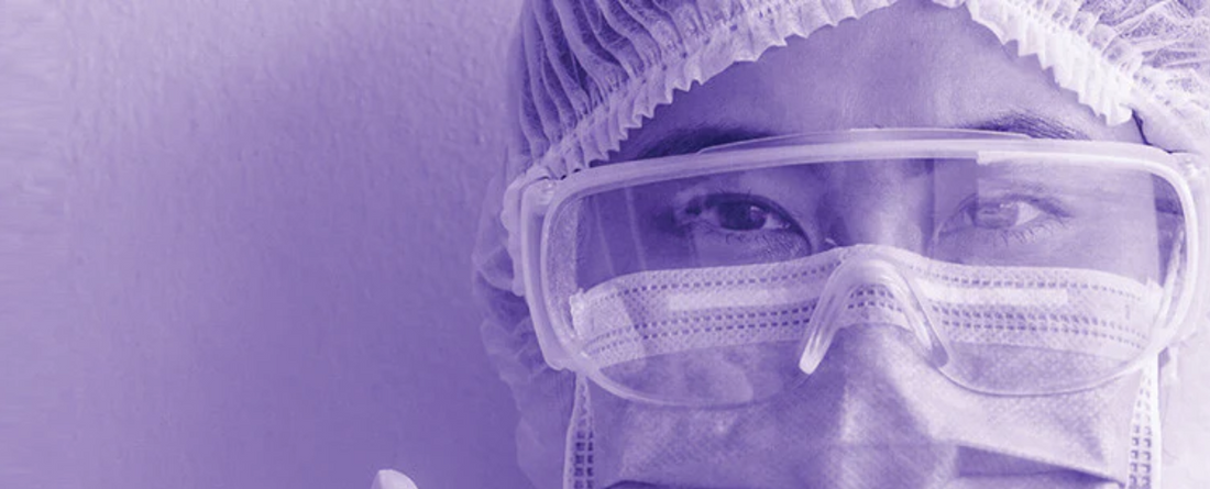 Healthcare worker wearing surgical mask, protective goggles, and hair cover in a purple-toned close-up