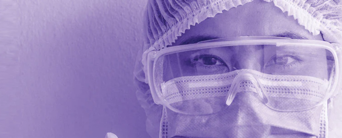 Healthcare worker wearing surgical mask, protective goggles, and hair cover in a purple-toned close-up
