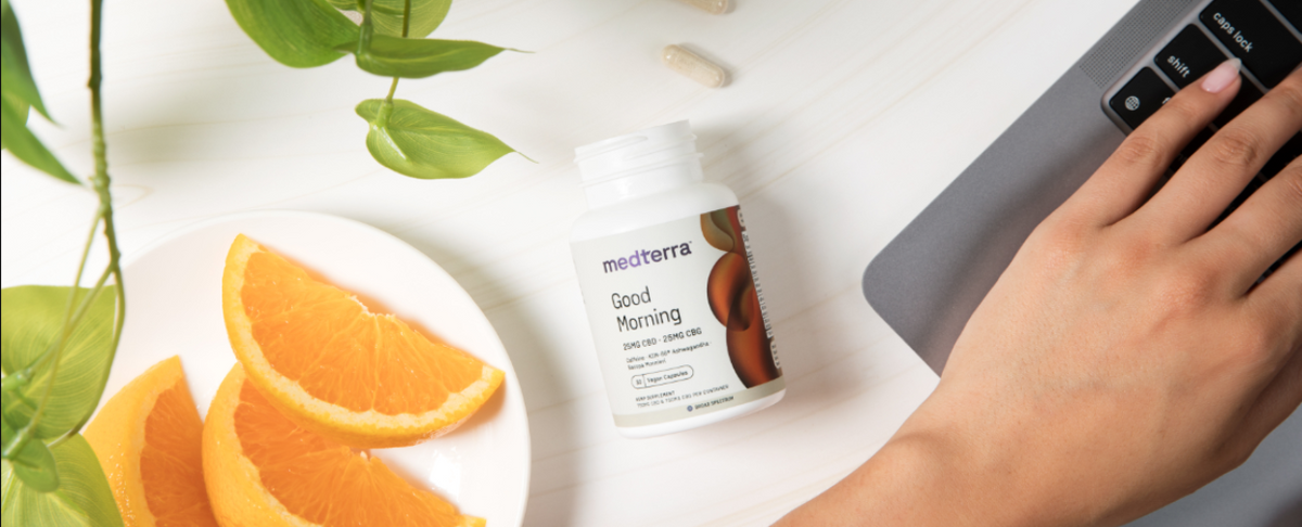 A hand reaching for a laptop next to a bottle of Medterra Good Morning capsules and orange slices on a plate.