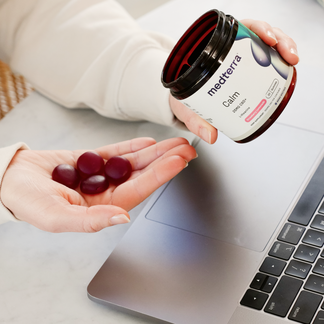 Hand holding four burgundy gummies beside a medterra jar labeled Calm 25MG CBD+ L-Theanine Strawberry Lemonade
