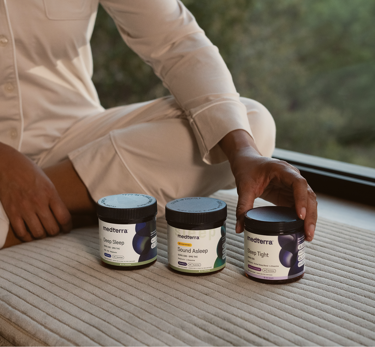 Three medterra jars on a bed labeled 'Deep Sleep', 'Sound Asleep', and 'Sleep Tight', with a hand reaching for one.