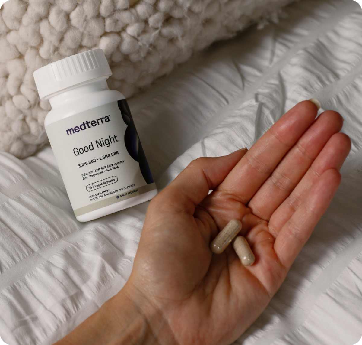 Angled close-up of Medterra Good Night CBD capsules bottle showing the label and two capsules held in a hand on white quilted bedding