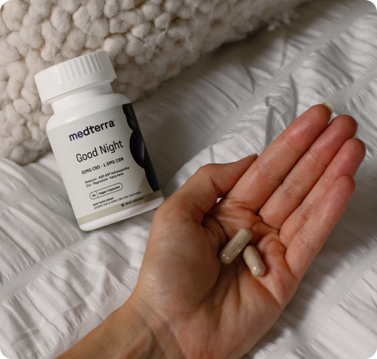 Close-up of Medterra Good Night capsule bottle with a hand holding two capsules on a white quilted bedspread