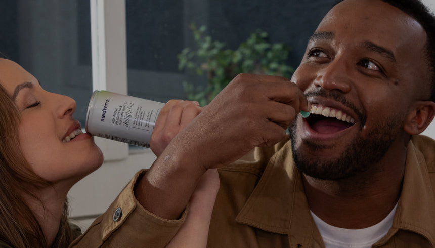 A smiling woman holds a cold beverage bottle for a man, who appears to be drinking from it. Both individuals are enjoying the moment in what appears to be a relaxed, indoor setting with a green plant and window visible in the background.