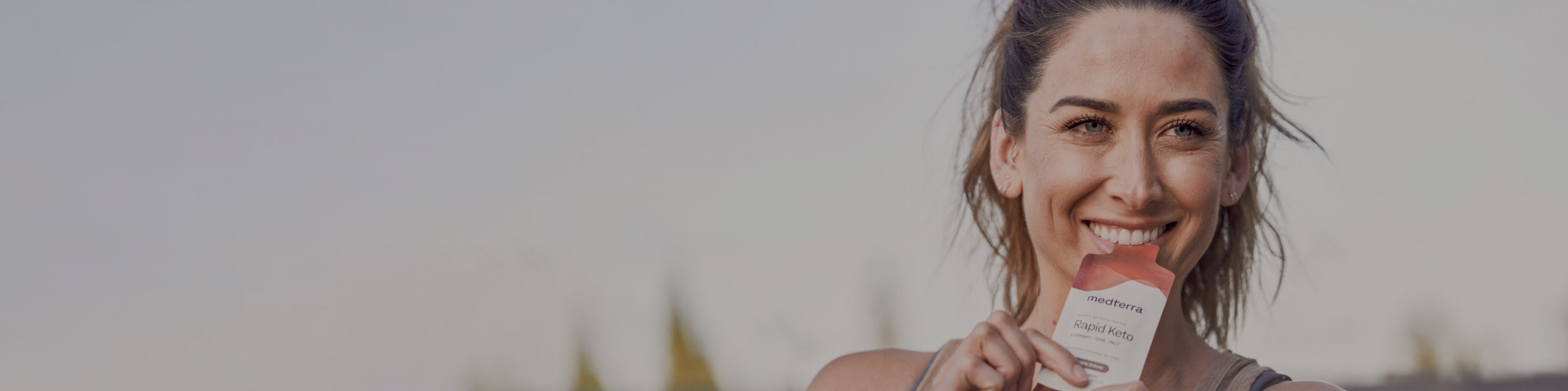 A smiling woman is holding a packaged product labeled "Medterra Rapid Keto" while standing outdoors. She has her hair tied back and is dressed in casual attire, with a blurred background of trees and a clear sky.