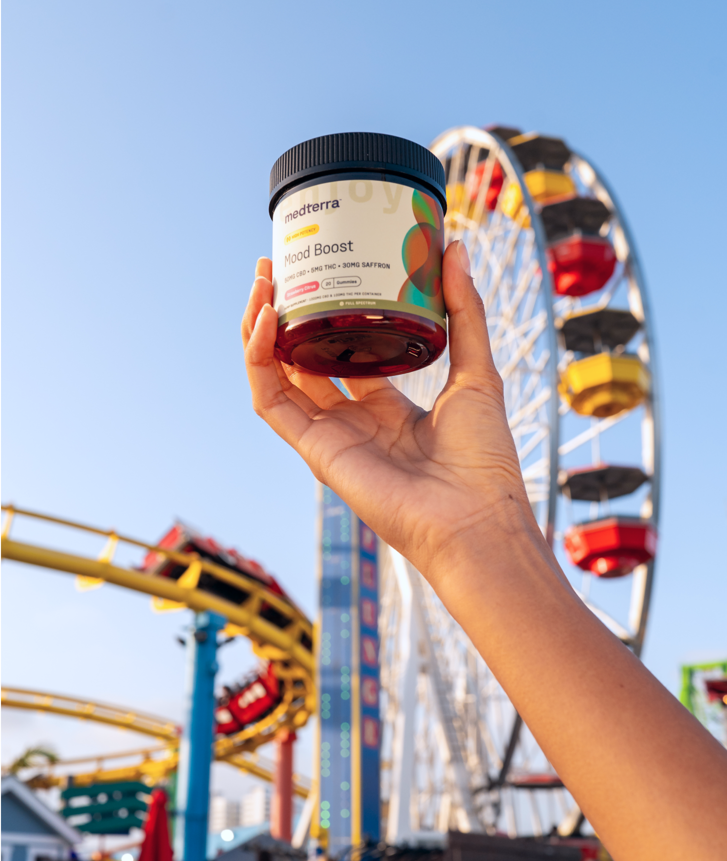 A hand is holding a Medterra Mood Boost supplement jar, made with the highest quality pure ingredients, with a colorful amusement park in the background featuring a ferris wheel and roller coaster against a clear blue sky.