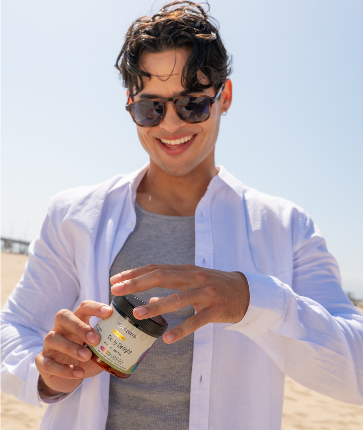 A smiling man wearing sunglasses and a white button-up shirt over a gray t-shirt stands on a sunny beach. He is holding a jar of Multivitamin Gummies made with pure ingredients and appears to be opening it. The sky is clear, and the beach is bright.