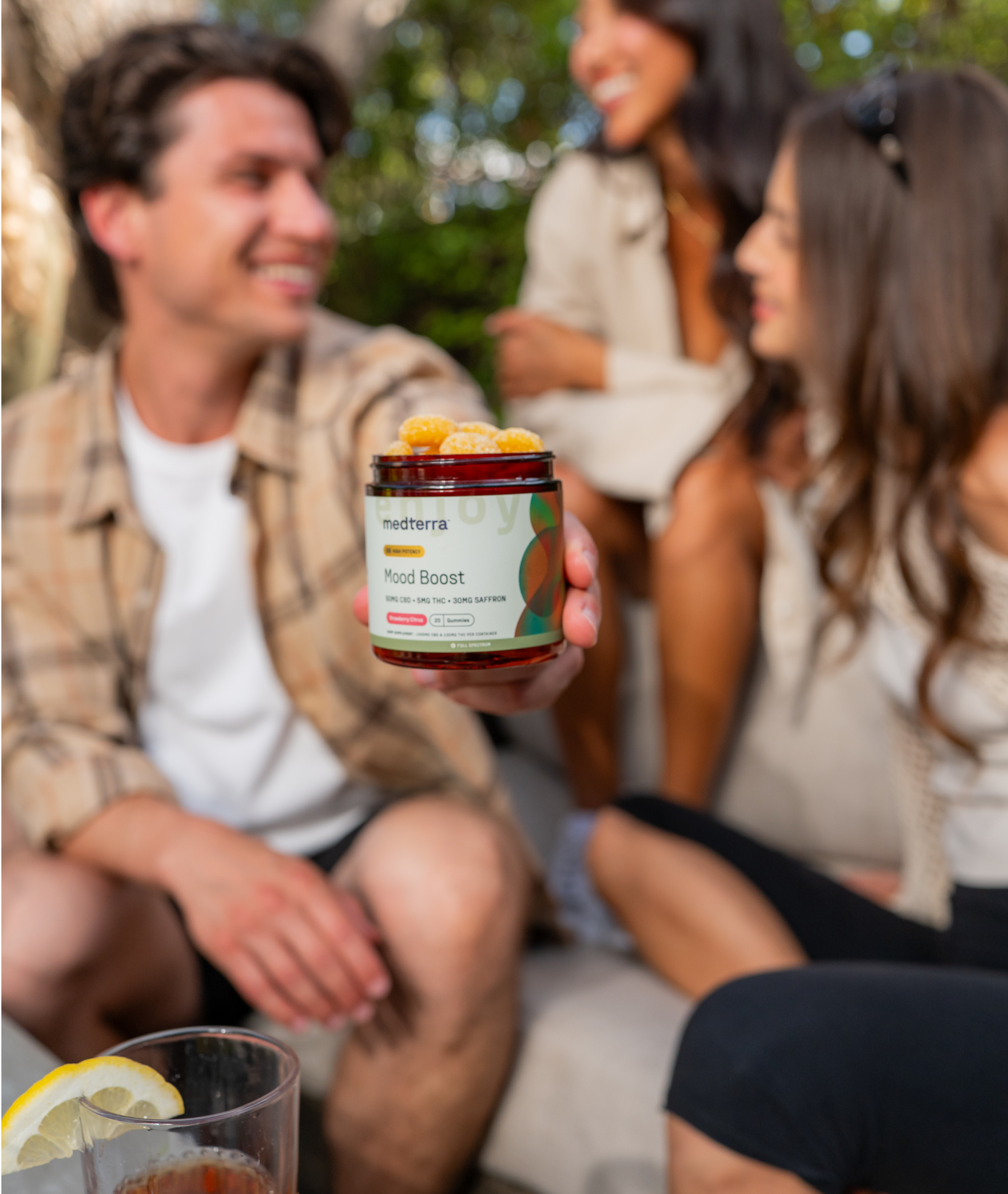 A smiling man holds a jar of Medterra Mood Boost gummies, made with the highest quality pure ingredients, towards the camera while sitting with two women. They appear to be in an outdoor setting, enjoying each other's company. The scene is bright and cheerful, with drinks on the table in the foreground.