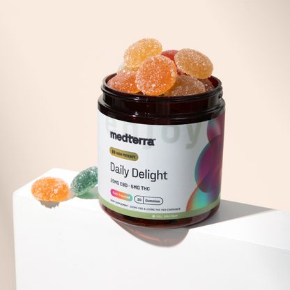 A jar labeled "Daily Delight Gummies" from Medterra contains colorful, sugar-coated wellness gummies. The label indicates each gummy has 25mg CBD and 5mg THC. The jar is placed on a white surface with a few gummies scattered around it. The text on the label reads, "High Potency.