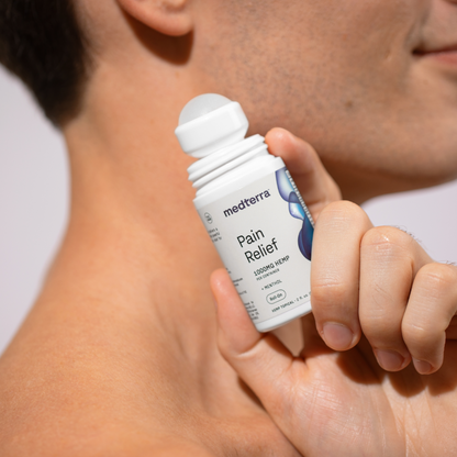 Close-up of a person applying Medterra Pain Relief Roll-On to their shoulder. The person's face is partially visible with a slight smile, and the focus is on the Pain Relief Roll-On by Medterra, featuring its 1000mg CBD formula, held up near the shoulder for joint pain relief.