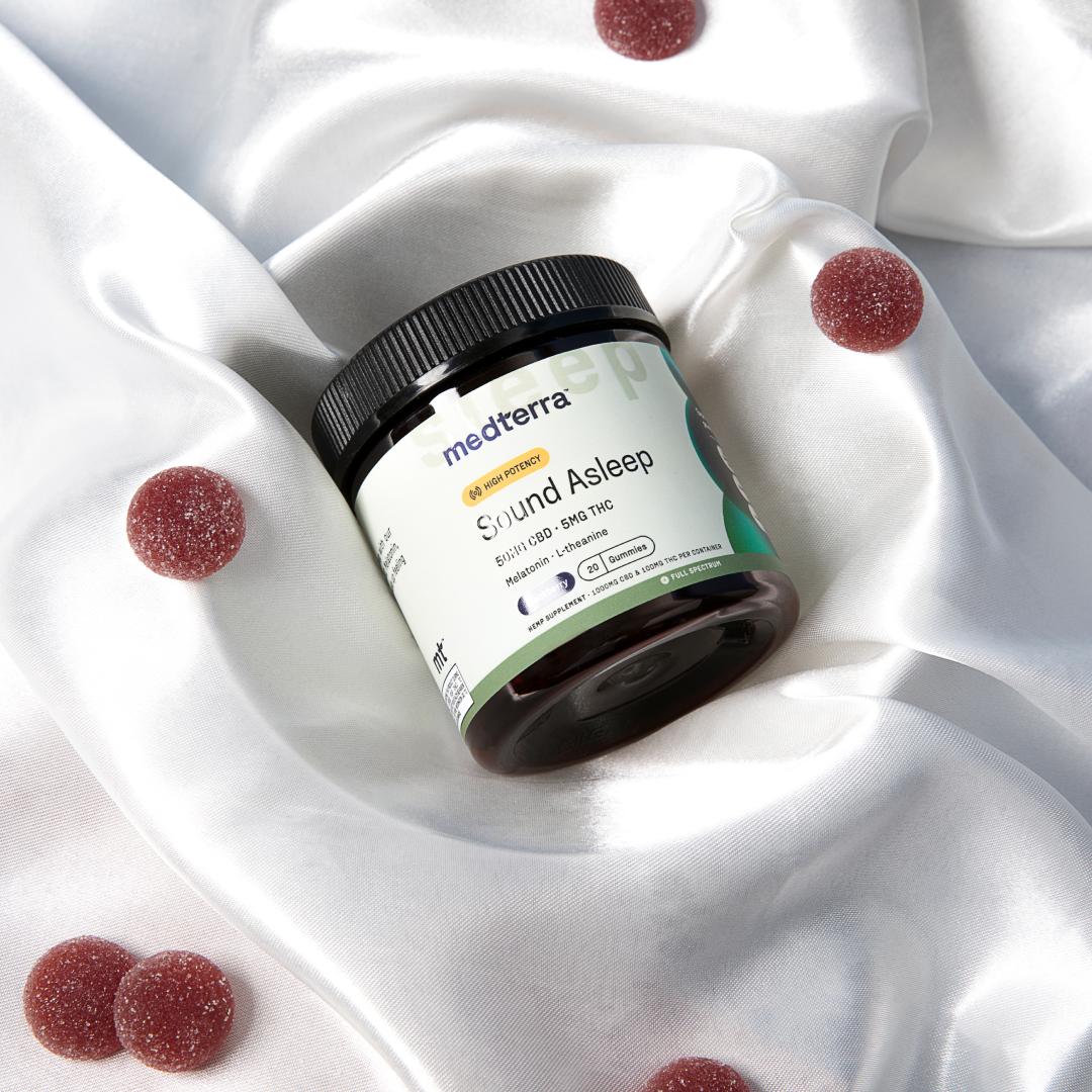 A container of Medterra brand CBD gummies labeled "Sound Asleep" rests on a silky white fabric. The container, surrounded by several round, red gummies scattered on the fabric, prominently lists THC-free details and melatonin dosage for its sleep aids.