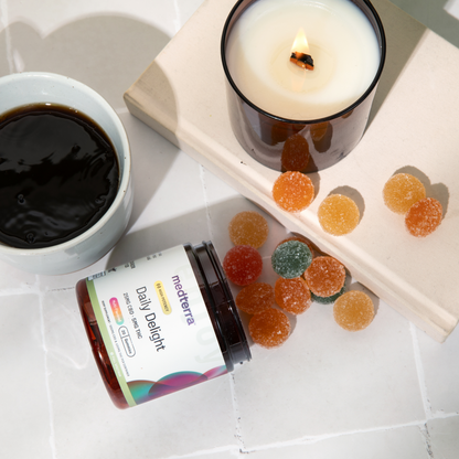 A jar of Medterra Daily Delight Gummies is open, with an assortment of gummies spilled out next to it. Beside the jar, there's a lit candle in a glass holder and a cup of black coffee on a white tile surface.
