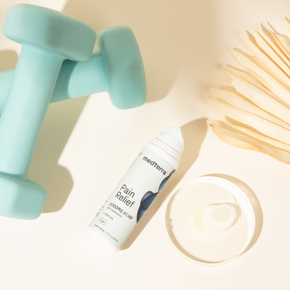 Two light blue dumbbells, a tube of Medterra Pain Relief Cream for arthritis and joint pain, and a round container of cream are arranged on a light surface beside some wooden sticks, likely for application.