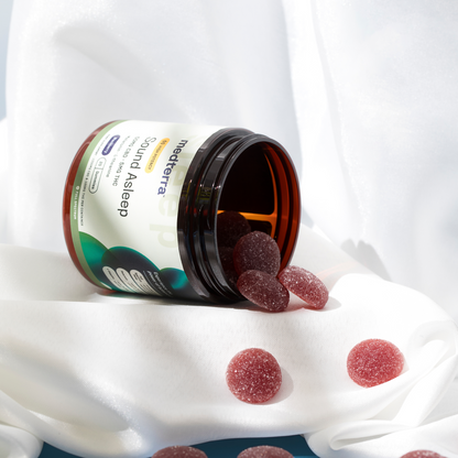 An open jar of Medterra Sound Asleep Gummies, a reliable sleep aid, with several dark red gummies spilling out onto a white cloth. The jar and gummies are softly illuminated, with a backdrop of white, flowing fabric.