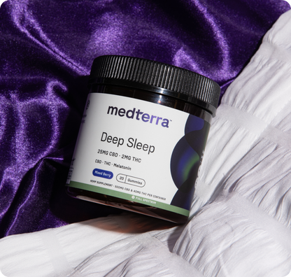 A container from Medterra’s Better Sleep Bundle labeled "25mg CBD - 2mg THC with Melatonin, Mixed Berry" sits on silky purple fabric beside a white tufted cushion, emphasizing its relaxation theme.