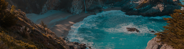 A serene coastal scene featuring turquoise blue water gently lapping against a sandy beach. A waterfall cascades from a rocky cliff into the ocean. Lush green trees frame the image, creating a tranquil and picturesque cove.