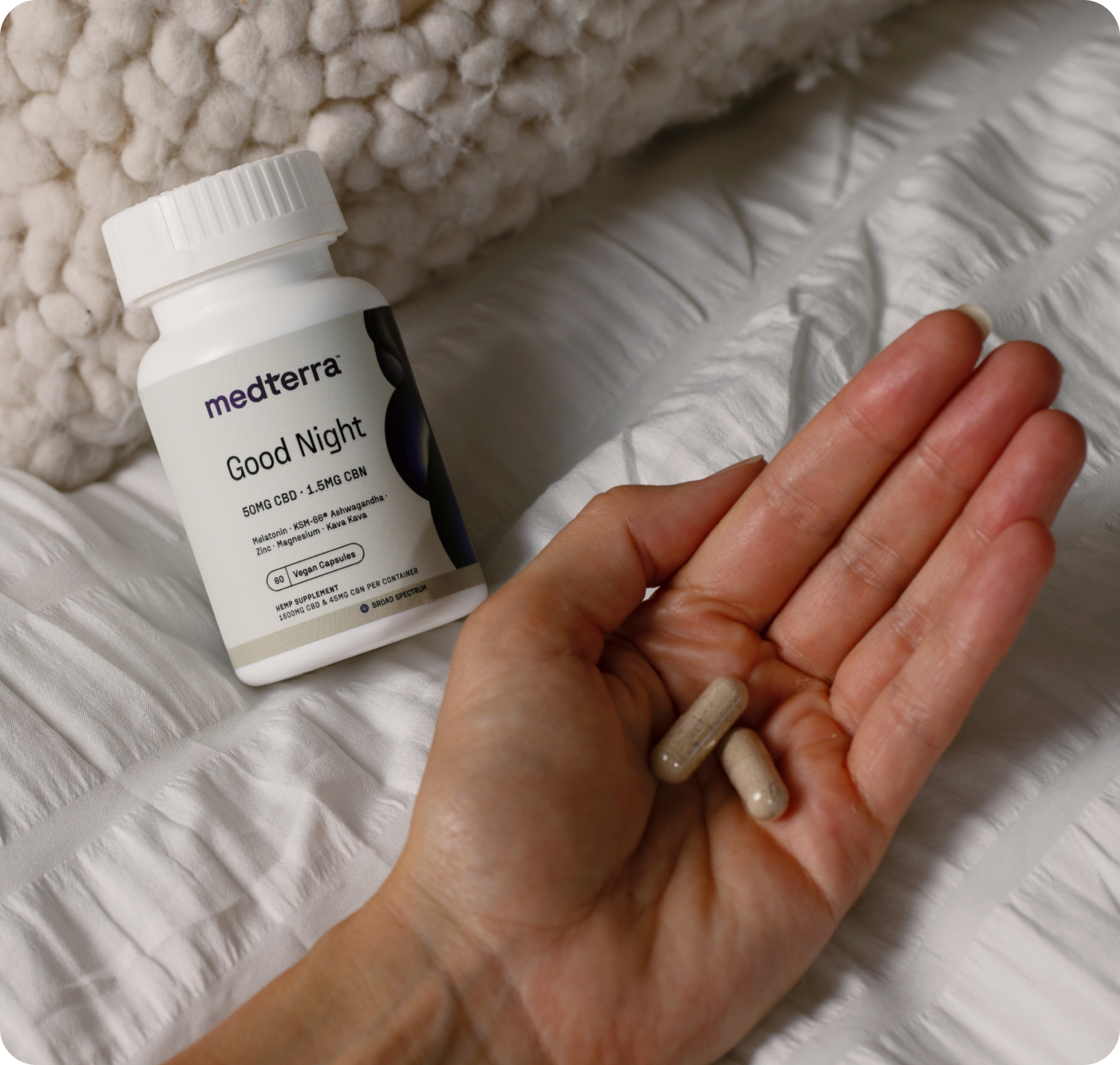 A hand holds two capsules over a white bedspread next to a Medterra Multi-Spectrum CBD Sleep Bundle bottle, with CBD and CBN content. A textured blanket is partially visible in the background.