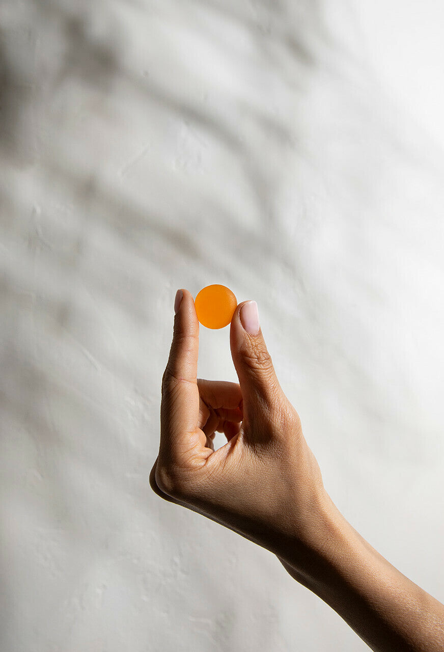 A close-up of a hand holding a small, round, amber-colored Max Relief Gummies [Free] from Medterra CBD between the thumb and index finger. The background is softly lit with a textured, neutral-colored surface, evoking the subtle elegance synonymous with premium Full Spectrum Hemp Extract products.