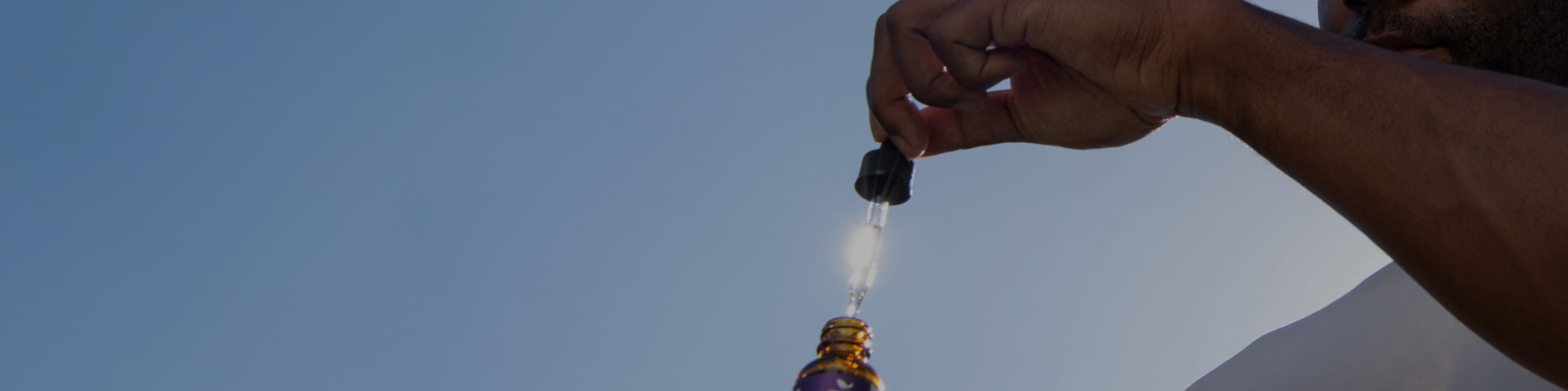 A person holds a dropper filled with liquid above a small, brown, glass bottle against a background of a clear blue sky. The close-up shot focuses on the dropper and bottle, with partial visibility of the person's hand and arm.