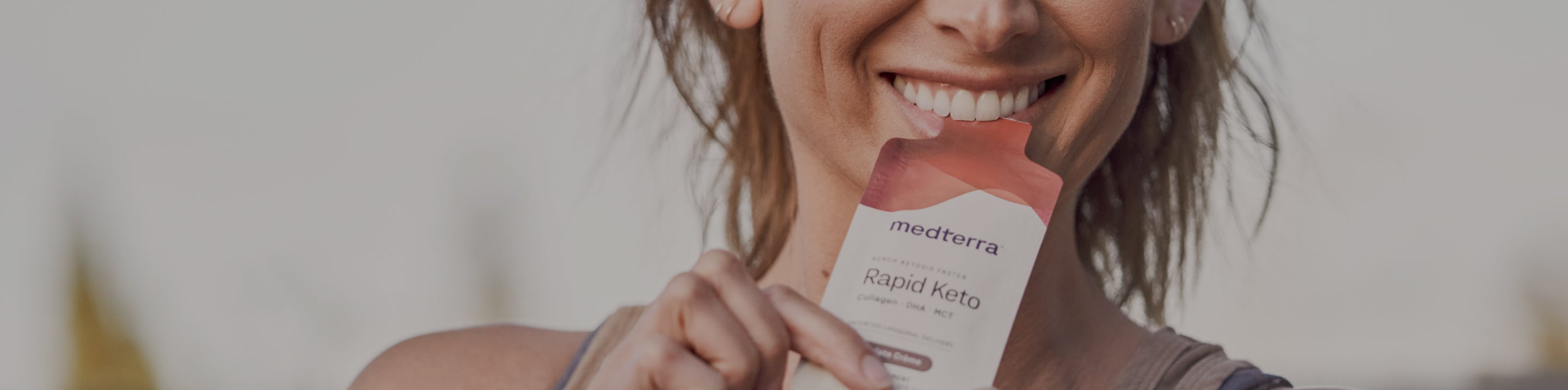 A person smiles while holding a "Medterra Rapid Keto" packet near their mouth. The focus is on the person’s mouth and the packet, highlighting the product. The background appears blurred, emphasizing the main subjects.