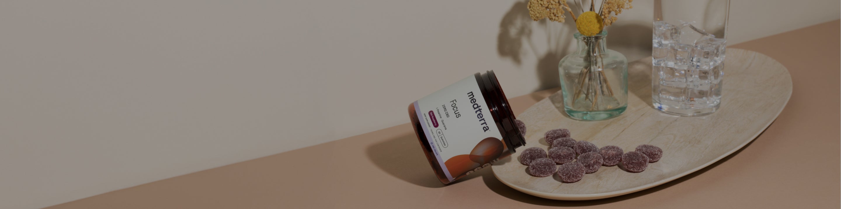 A jar of Medterra Focus gummies lies on its side, spilling gummies onto a wooden tray. Beside the tray are two glass vases with flowers and a glass of water. The background is neutral with a light beige wall and soft peach surface.