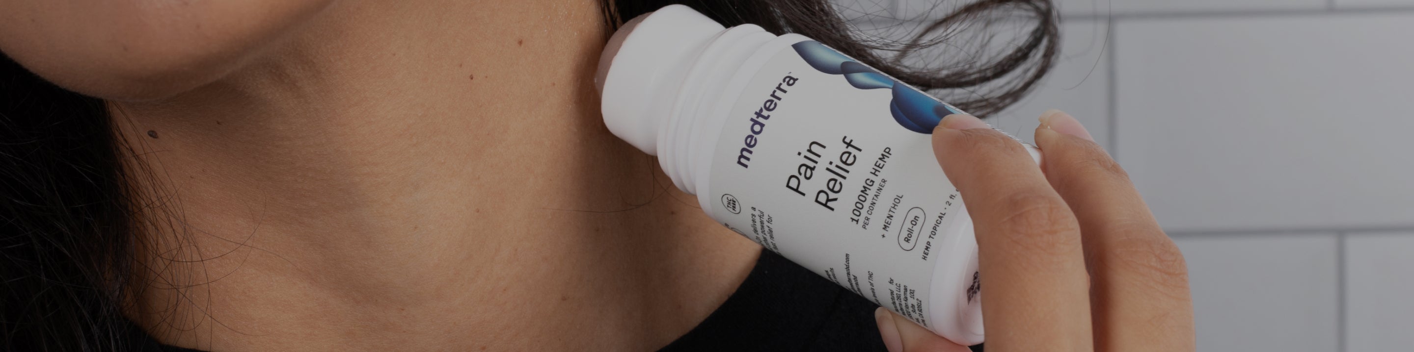 A person applying Medterra Pain Relief Cream with a roll-on applicator to the side of their neck. The label on the container is partially visible, showing the brand and some product details. The background is blurred, focusing attention on the application.