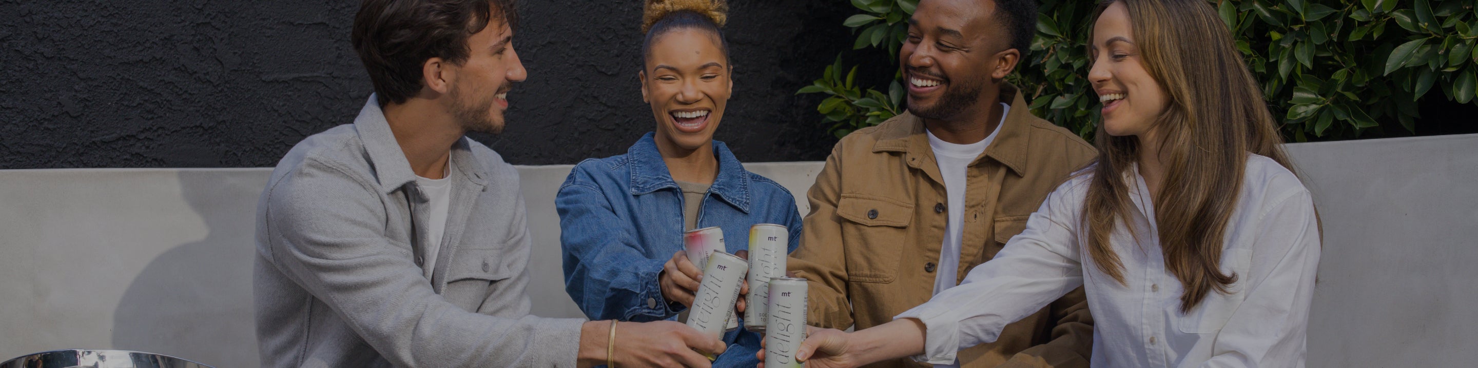 A group of four people, two men and two women, sit on a bench outdoors, smiling and clinking Medterra Delight Social Tonic cans together as they celebrate. They are casually dressed and appear to be enjoying a friendly, relaxed gathering. Green foliage is visible in the background.