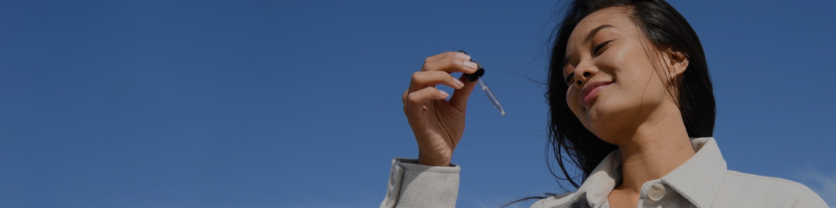 A woman with long dark hair holds a Medterra tincture dropper up to the sky with a smile on her face. She is wearing a light-colored jacket, and the background is a clear blue sky.