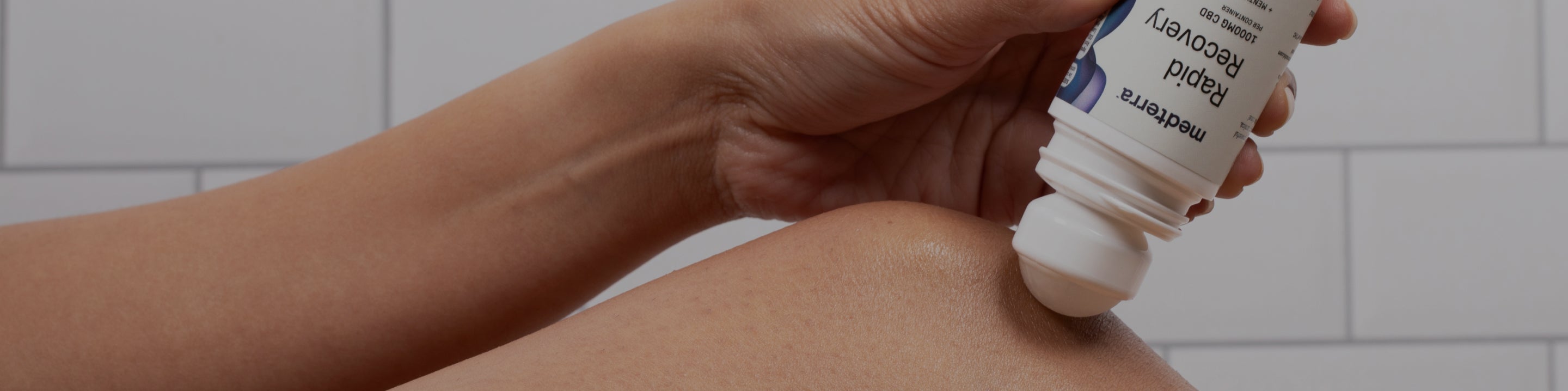 A person's hand is applying a roll-on product labeled "Rapid Recovery" to their knee. The background features white tiled walls.