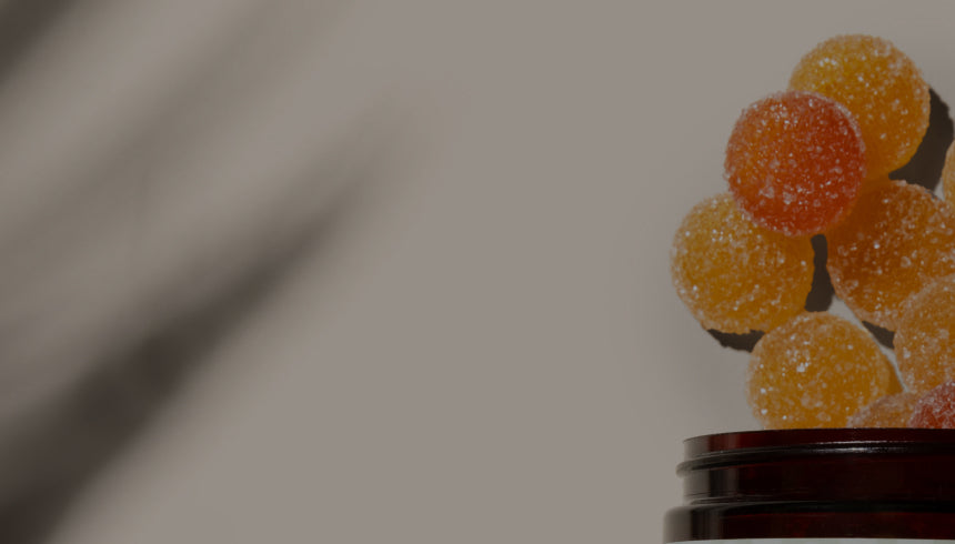 Round, Medterra gummies in various shades of orange and red are seen in mid-air above a brown jar, as if they are being poured out or are floating. The background is blurred gray, and the candies appear vibrant against it.