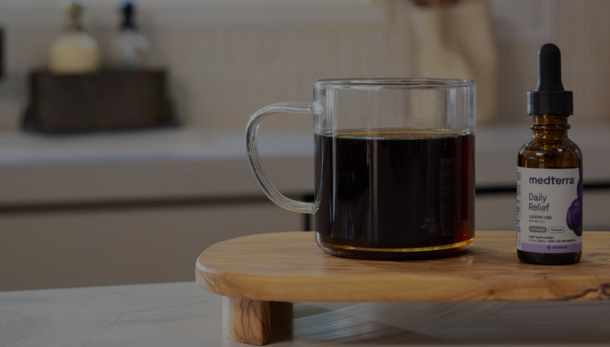 A clear glass cup filled with dark liquid is next to a small brown glass dropper bottle labeled "daily relief" on a wooden platform. The background features a modern kitchen countertop with various items out of focus.