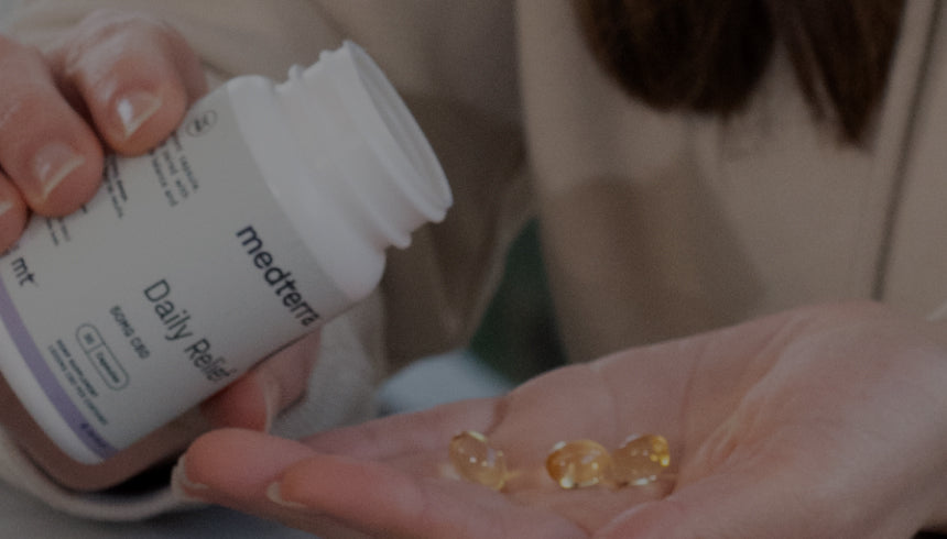 A person holding a white container labeled "Medterra Daily Relief" in one hand and pouring out several yellow capsules into the other hand. The background shows a blurred workspace with a laptop and plant.