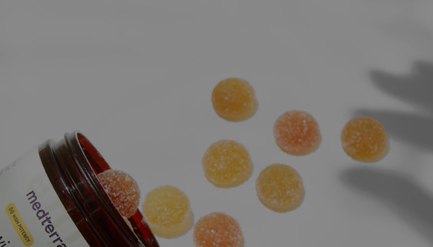 A brown jar is tipped over, spilling several round, sugar-coated gummies onto a white surface. A hand's shadow reaches towards the scattered gummies, indicating someone about to pick them up.