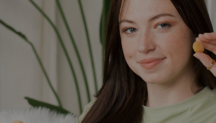 A woman with long brown hair and light skin holds a Medterra gummy near her face. She is smiling slightly. In the background, there are green, leafy plants.