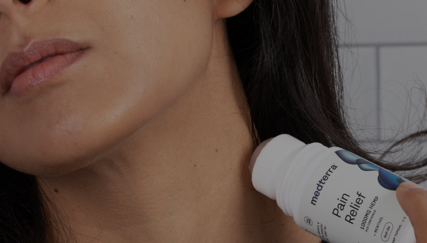 A person applying Medterra Pain Relief Cream with a roll-on applicator to the side of their neck. The label on the container is partially visible, showing the brand and some product details. The background is blurred, focusing attention on the application.