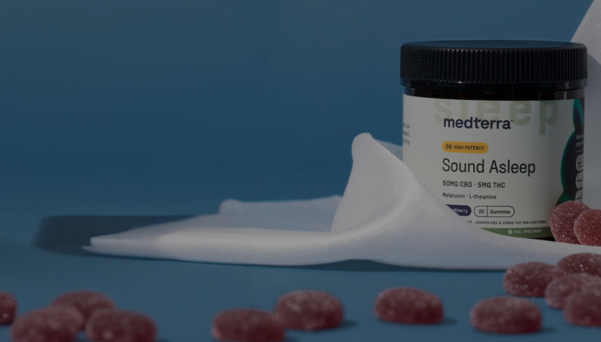 A jar of Medterra Sound Asleep CBD gummies is placed against a blue background with a white cloth draped around it. The jar is labeled with product information, and several red gummies are scattered on the blue surface in the foreground.