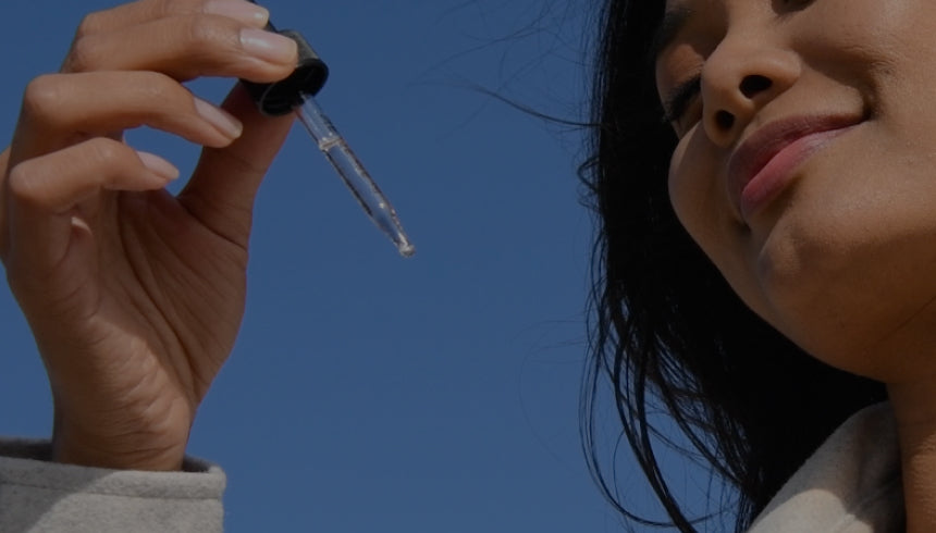 A woman with long dark hair holds a Medterra tincture dropper up to the sky with a smile on her face. She is wearing a light-colored jacket, and the background is a clear blue sky.