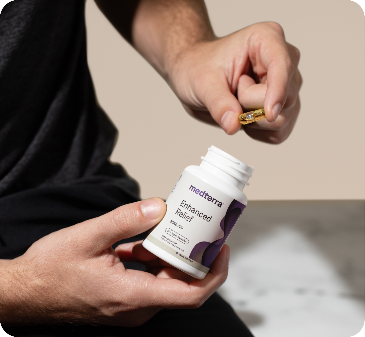 A person holding a CBD pill above an open bottle labeled "medterra Enhanced Relief" with their other hand. The person is wearing a dark shirt and the background is blurred, showing a marble surface, suggesting this might be the solution to relieve stress and elevate your mood.
