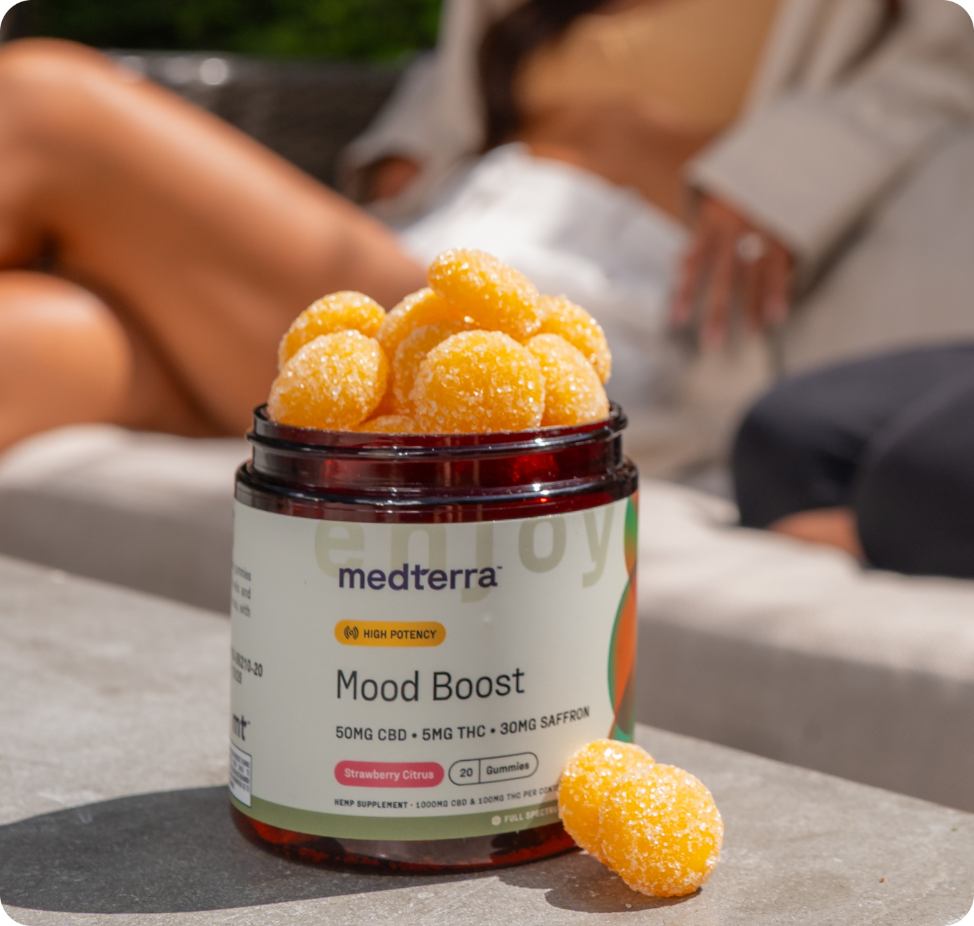 On a table sits a jar from Medterra's Mother's Day Bundle, labeled "Mood Boost," containing orange gummies. The label shows "50mg CBD," "5mg THC," and "30mg Saffron." In the background, two people relax on an outdoor couch.