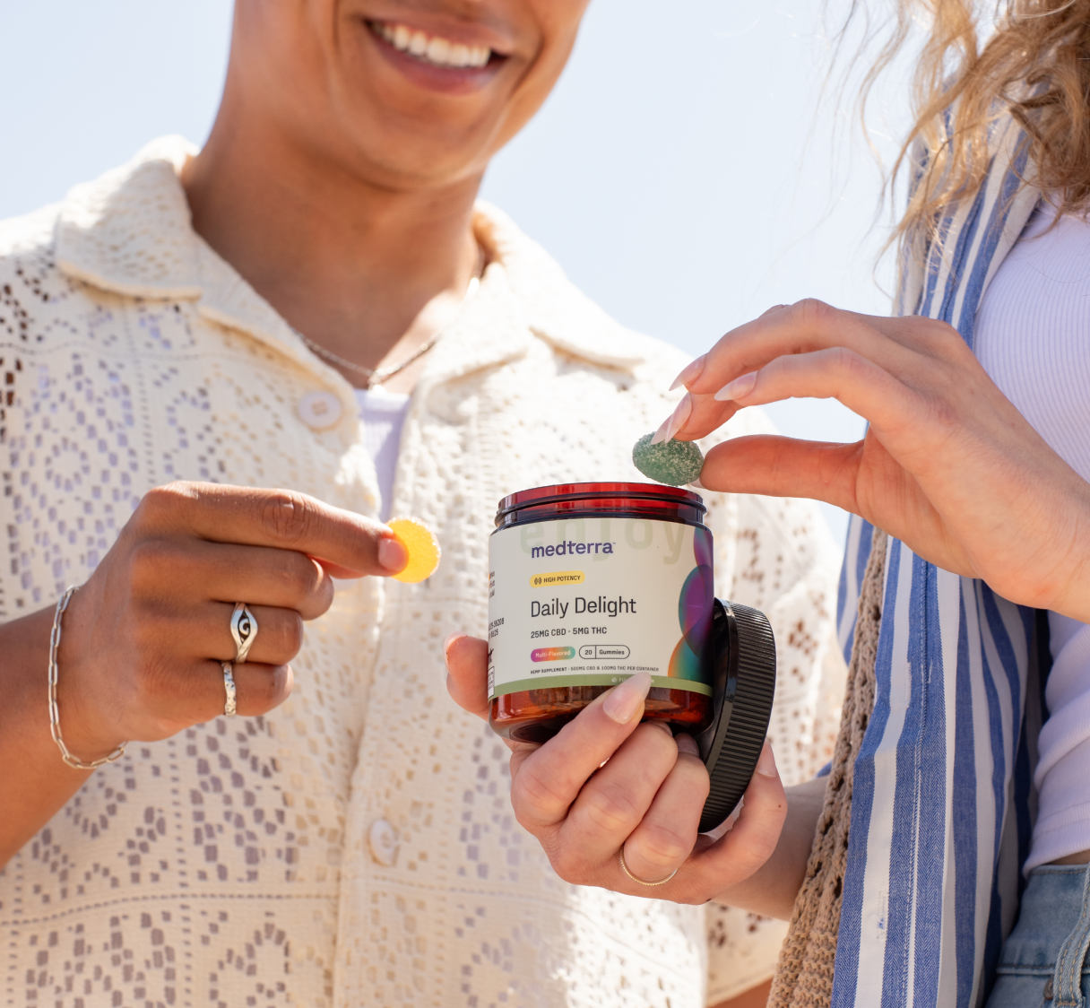 Two people are holding a jar of Medterra Daily Delight CBD gummies. One person is holding a yellow gummy, while the other reaches for a green gummy from the jar. The background is bright and sunny, highlighting their smiles as they enjoy quality CBD.