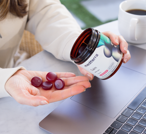 A person is holding a jar of Medterra Calm CBD gummies, with several red gummies in hand. The jar label, indicating it's Ultra Broad Spectrum, is clearly visible. The person is sitting at a table with a laptop and a cup of coffee beside them.