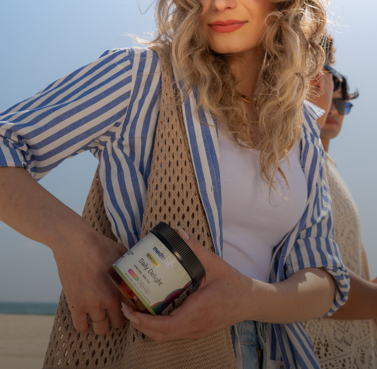 A woman with wavy blonde hair, wearing a white shirt and blue-striped cardigan, holds a jar of MaryRuth Organics Daily Greens. She's outdoors on a sunny day, standing next to another person, with a beach and blue sky in the background. The scene radiates calmness and tranquility.