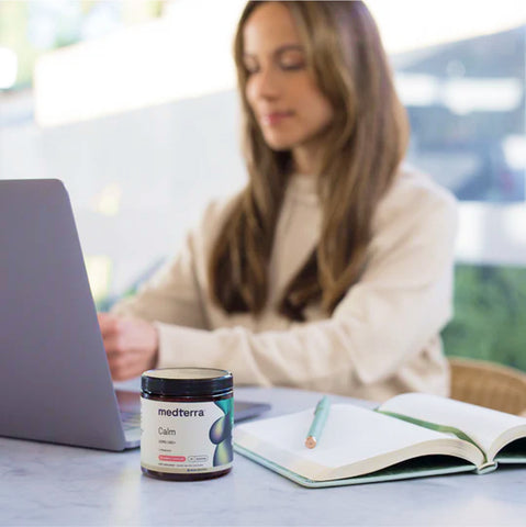 A woman sits at a desk working on her laptop, with a notebook and a pencil next to her. In the foreground, there is a jar labeled "Medterra Calm," promoting high-quality CBD products. The background is softly blurred, emphasizing the woman and her workspace. 