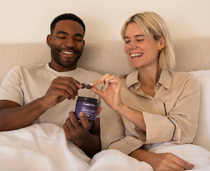 A smiling couple sits together on a bed, sharing a moment with a jar labeled "Sleep Tight Gummies [Top 10]." The man holds the jar of premium Medterra CBD Sleep Tight Gummies and offers one to the woman, who is reaching out to take it. Both appear relaxed and happy.